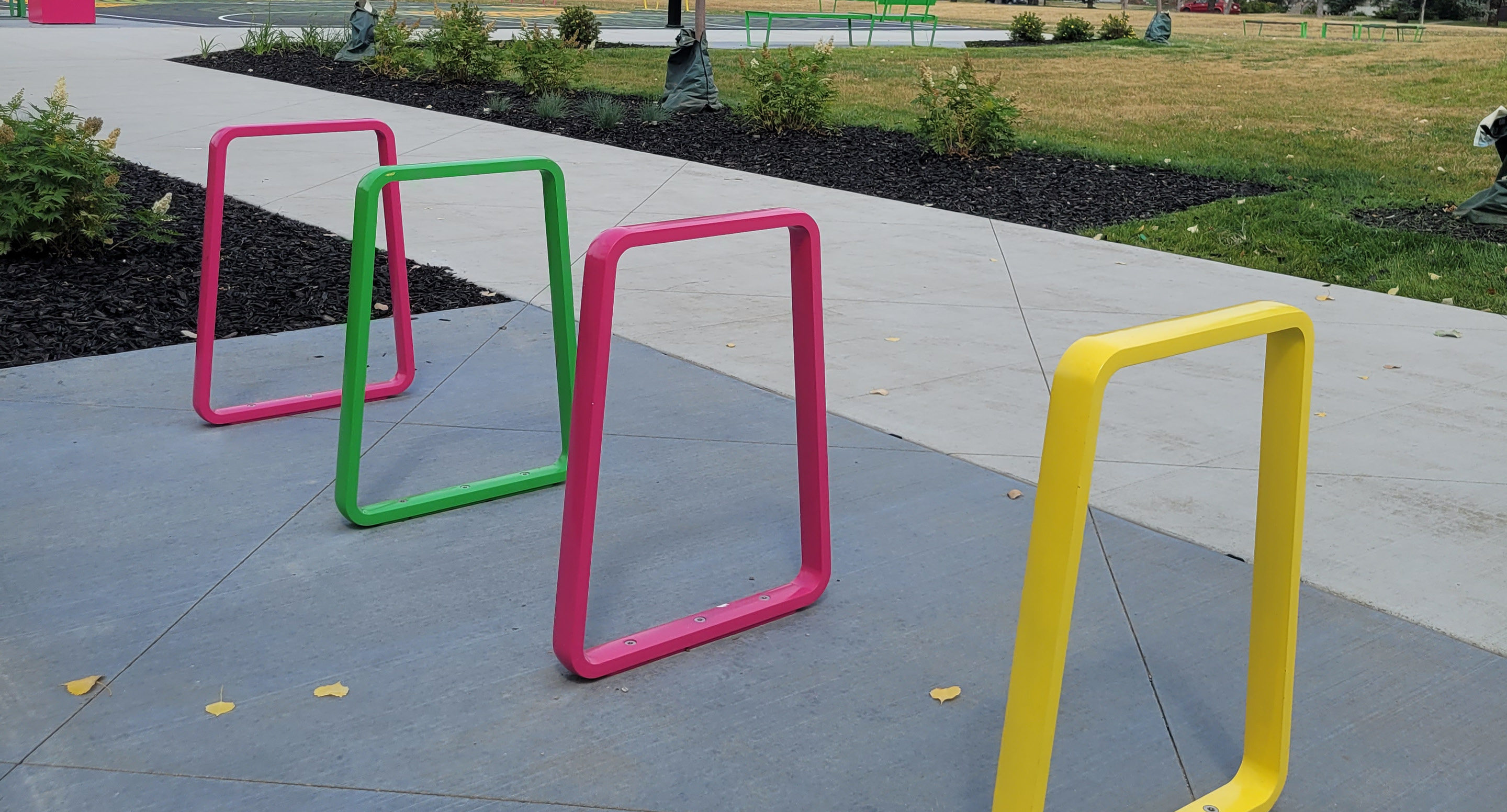 maglin-collections-iconic_gallery8 Three colorful metal bike racks—pink, green, and yellow—are installed on a sidewalk in an outdoor park area with grass, shrubs, and a basketball court in the background.