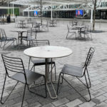 Several empty metal tables and chairs are arranged on a paved outdoor plaza with some leafless trees and modern buildings in the background.