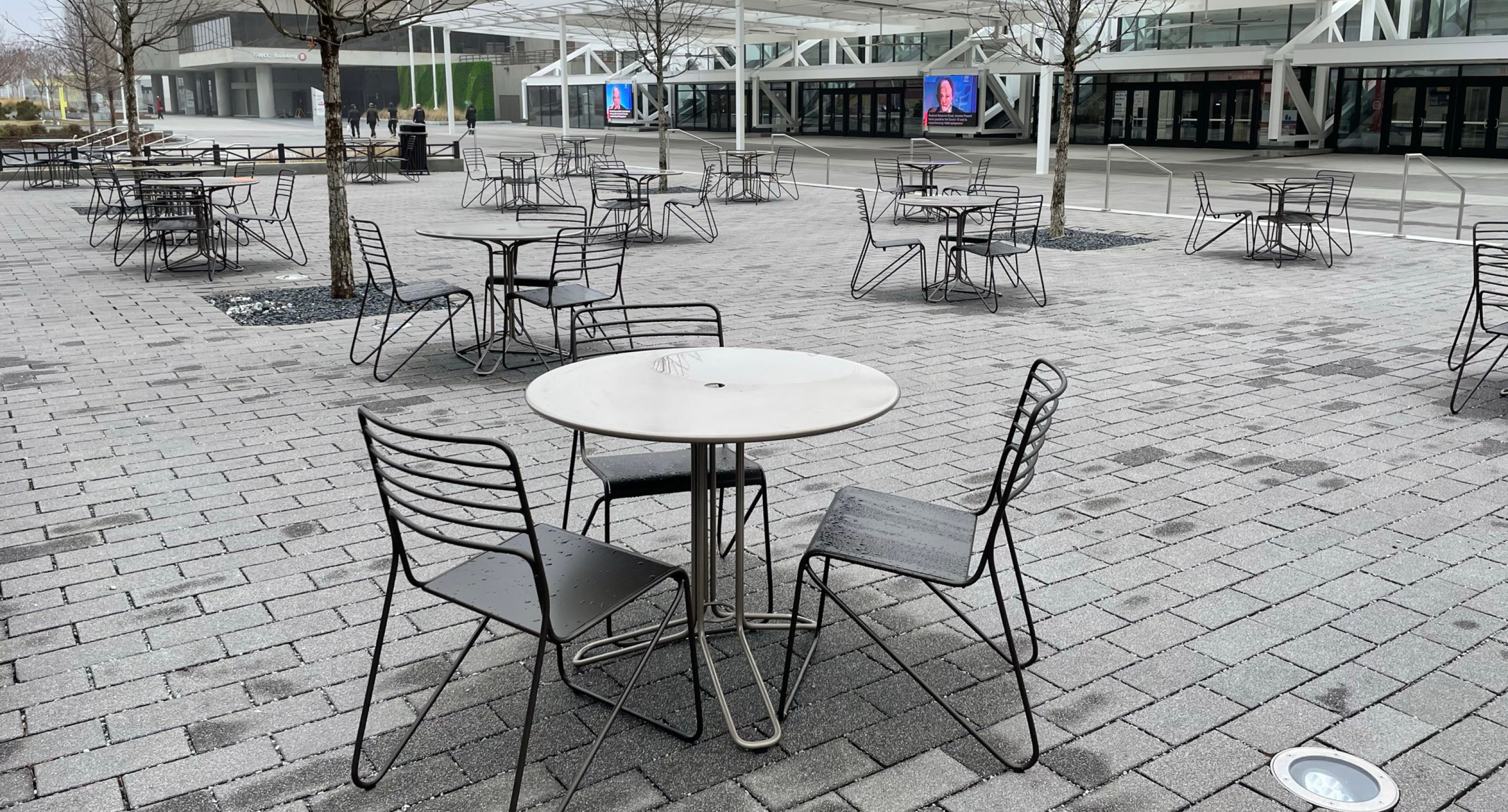 Several empty metal tables and chairs are arranged on a paved outdoor plaza with some leafless trees and modern buildings in the background.