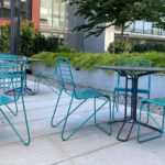 Several empty turquoise metal chairs and tables are arranged on a concrete patio outdoors, with modern buildings and greenery in the background.