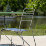 A metal chair and green table are set on a concrete patio by a pond, with trees and greenery in the background.