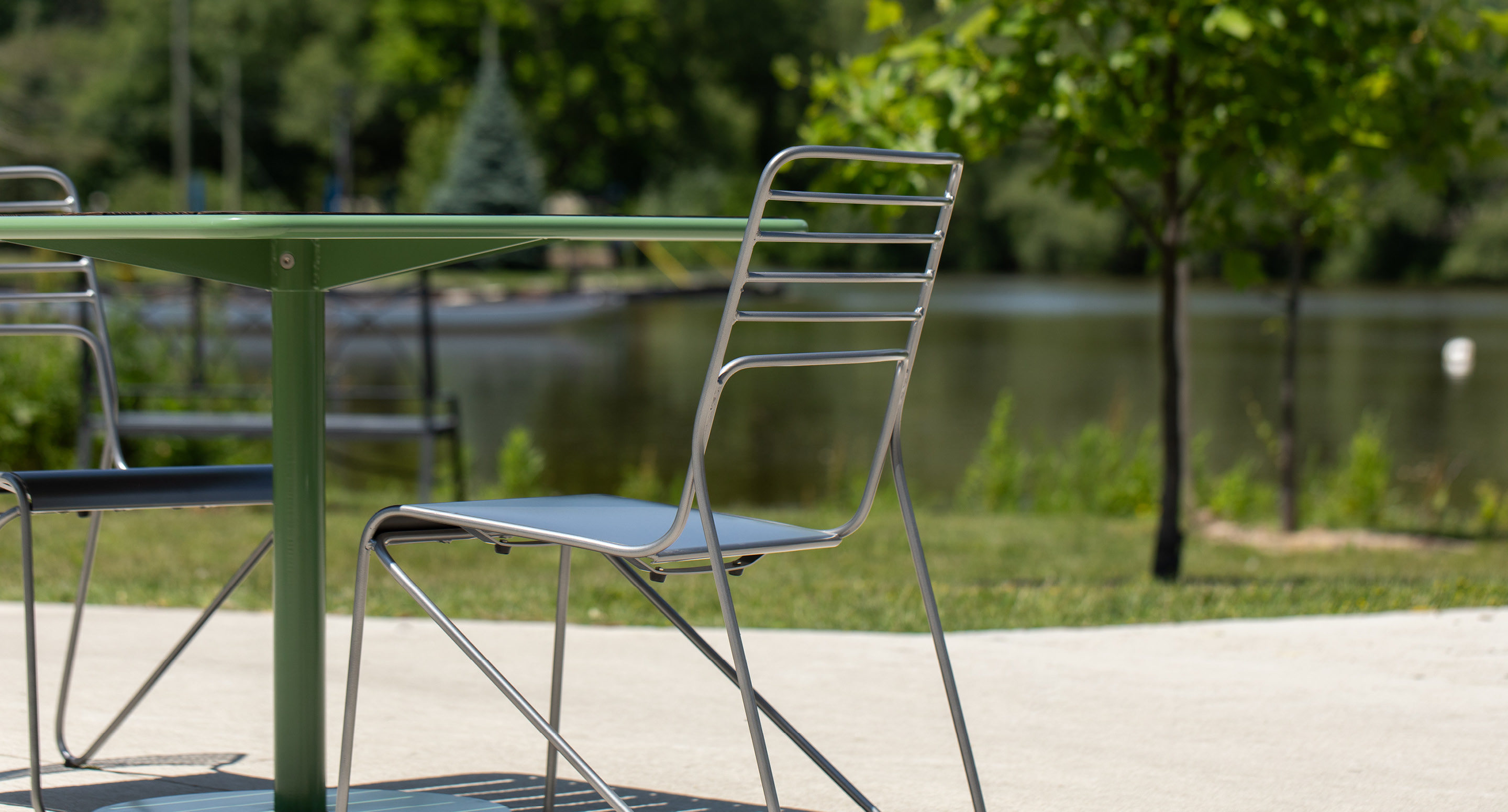 maglin-collections-kontur-gallery4 A metal chair and green table are set on a concrete patio by a pond, with trees and greenery in the background.