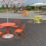 Outdoor seating area with orange and yellow metal tables and chairs, large umbrellas, and paved walkways in a park setting with greenery and sports equipment in the background.