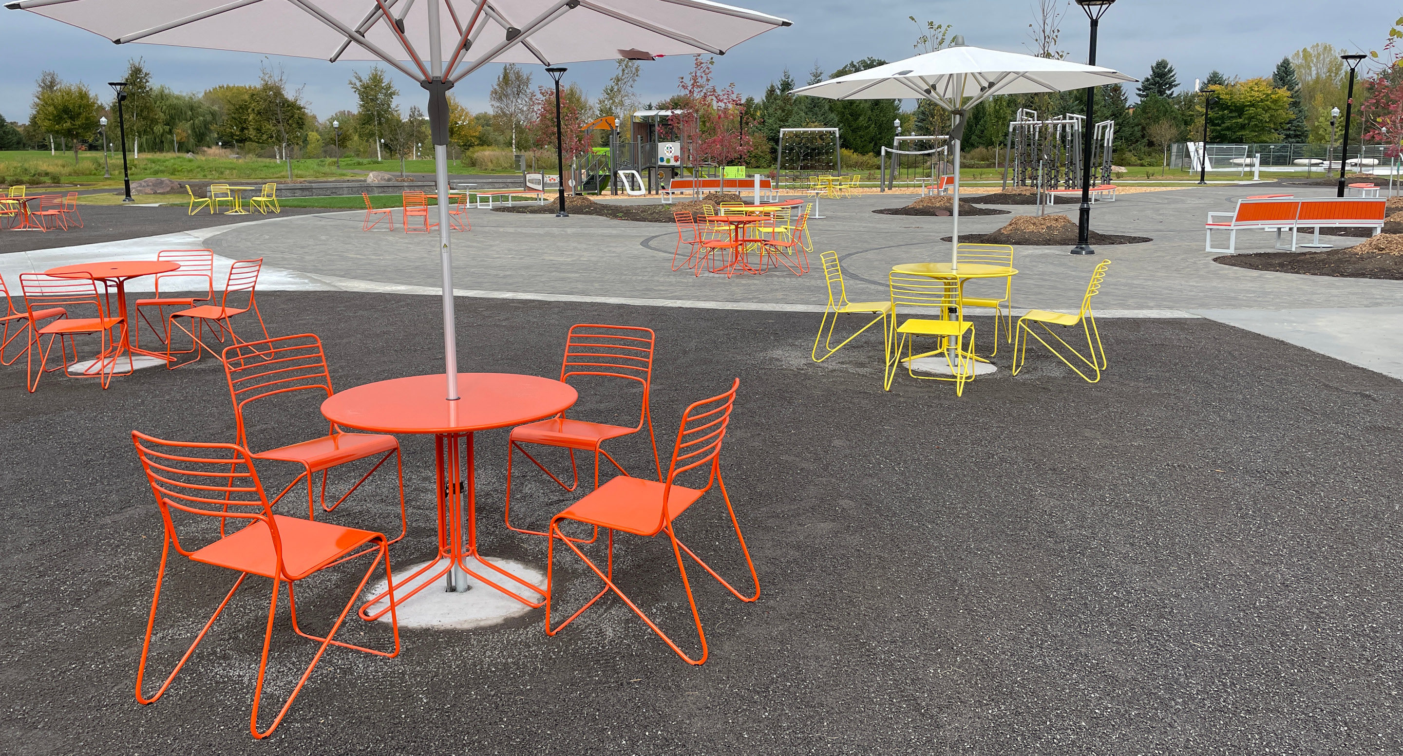 maglin-collections-kontur-gallery5 Outdoor seating area with orange and yellow metal tables and chairs, large umbrellas, and paved walkways in a park setting with greenery and sports equipment in the background.