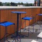 A small blue metal table with three matching blue stools is placed on a tiled outdoor patio with wooden paneling and a partial city view in the background.