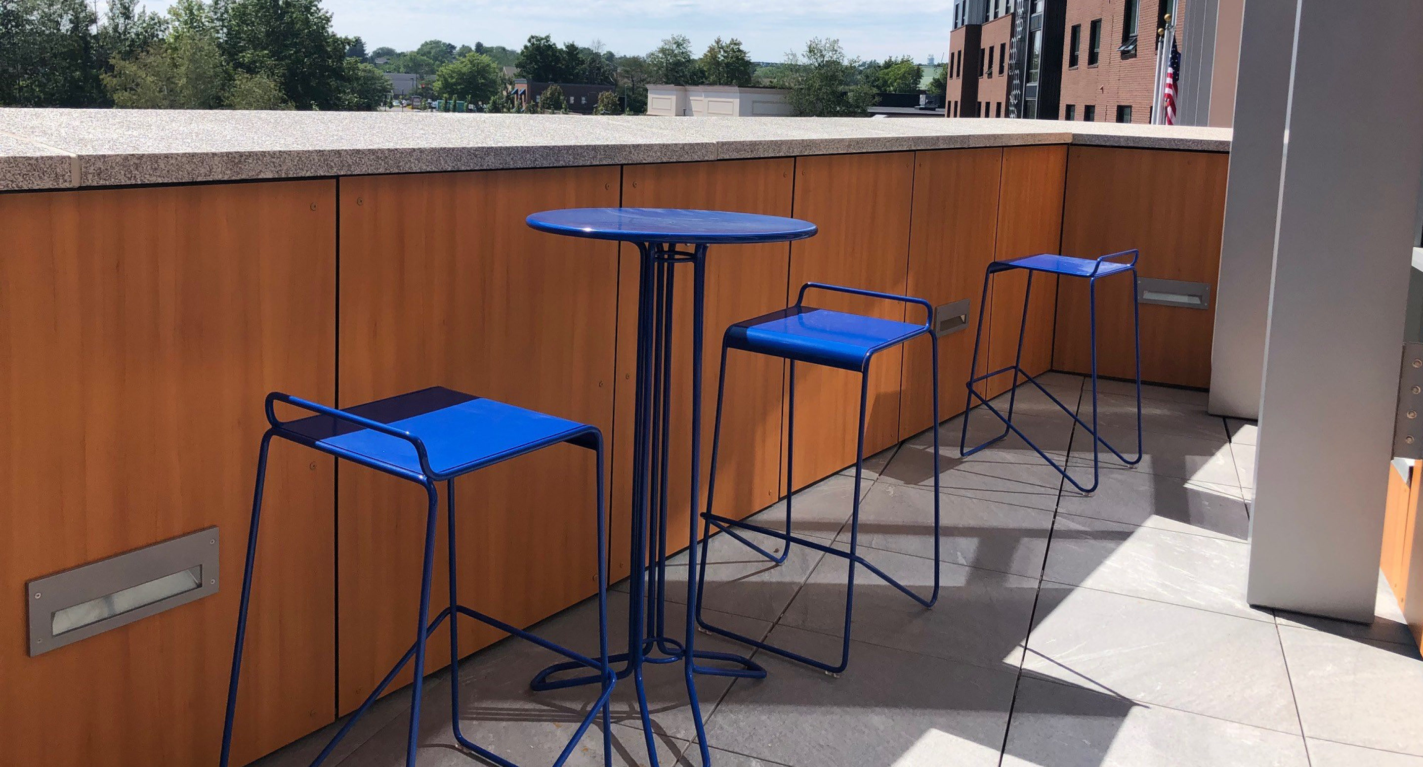 A small blue metal table with three matching blue stools is placed on a tiled outdoor patio with wooden paneling and a partial city view in the background.