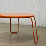 A small round orange metal table with curved legs stands on a polished concrete floor in front of a white textured wall.