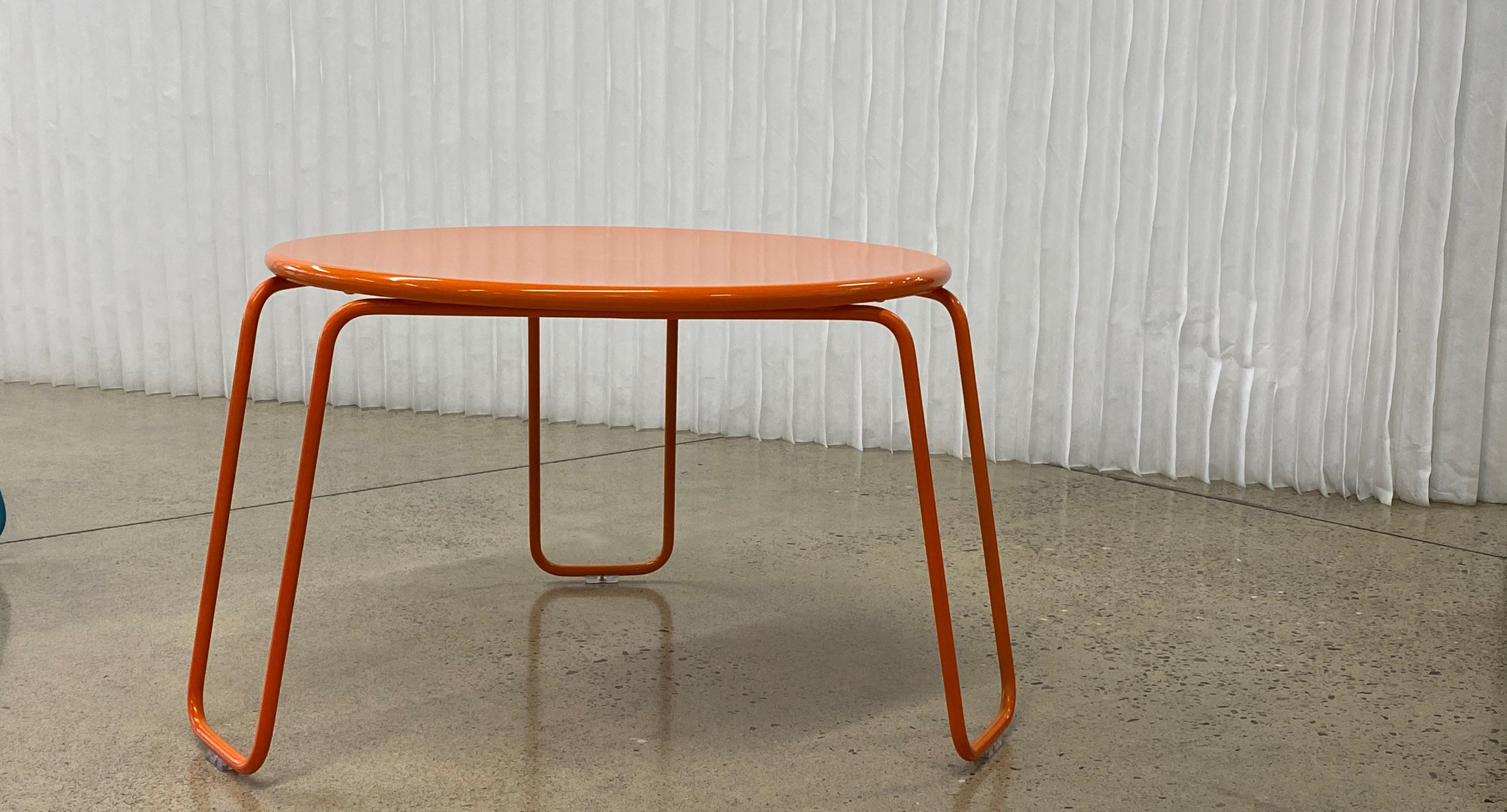 maglin-collections-kontur-gallery7 A small round orange metal table with curved legs stands on a polished concrete floor in front of a white textured wall.