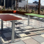 Two rectangular tables, one red and one green, with metal chairs on a tiled rooftop terrace under partial shade, with city buildings in the background.
