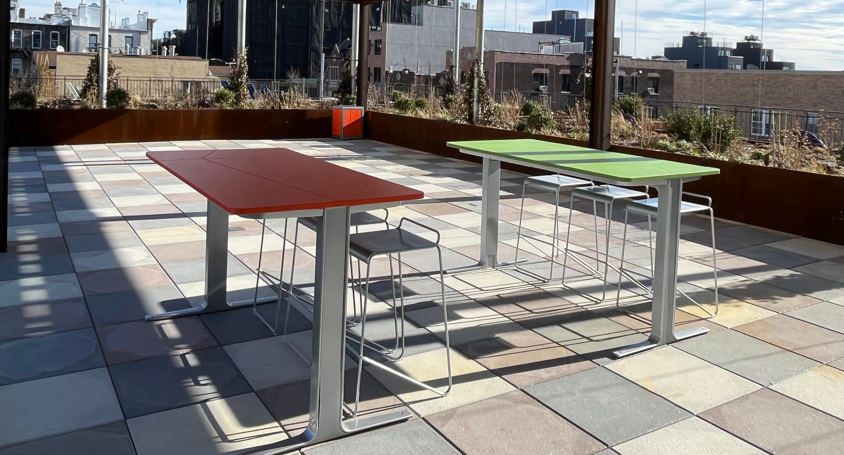 Two rectangular tables, one red and one green, with metal chairs on a tiled rooftop terrace under partial shade, with city buildings in the background.