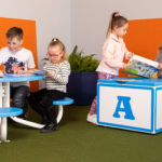 Quatre jeunes enfants sont assis à une table et lisent des livres, tandis que deux filles se tiennent près d'un grand bloc bleu et blanc, l'une d'elles tenant un livre d'images ouvert. La pièce a des cloisons orange et un sol vert.