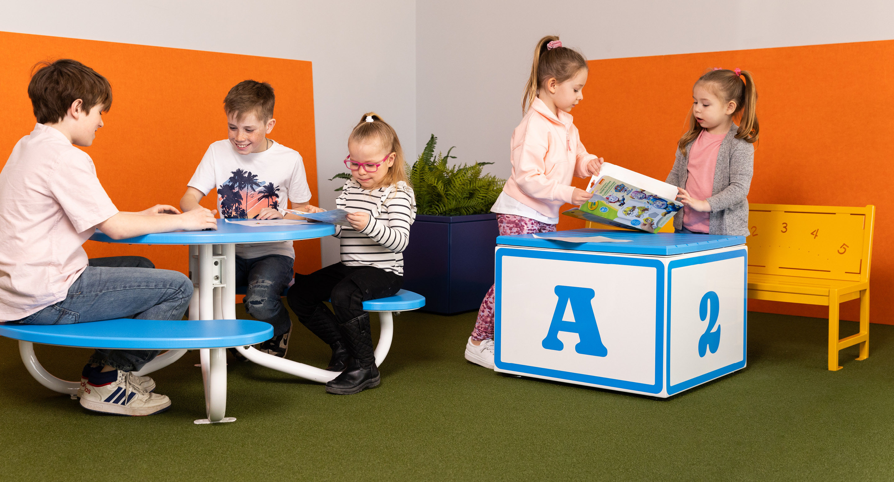 maglin-collections-minis-hero Quatre jeunes enfants sont assis à une table et lisent des livres, tandis que deux filles se tiennent près d'un grand bloc bleu et blanc, l'une d'elles tenant un livre d'images ouvert. La pièce a des cloisons orange et un sol vert.
