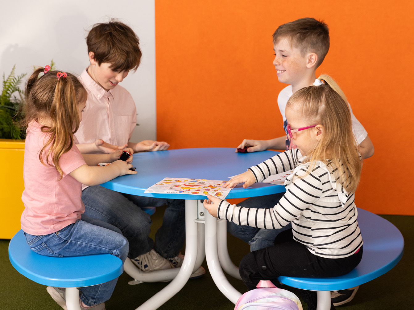 Four children sit around a blue circular table, playing and looking at a colorful activity sheet together in a bright room.