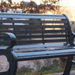 A black metal bench is positioned outdoors on a paved walkway, with a blurred pedestrian and ornamental grasses in the background.