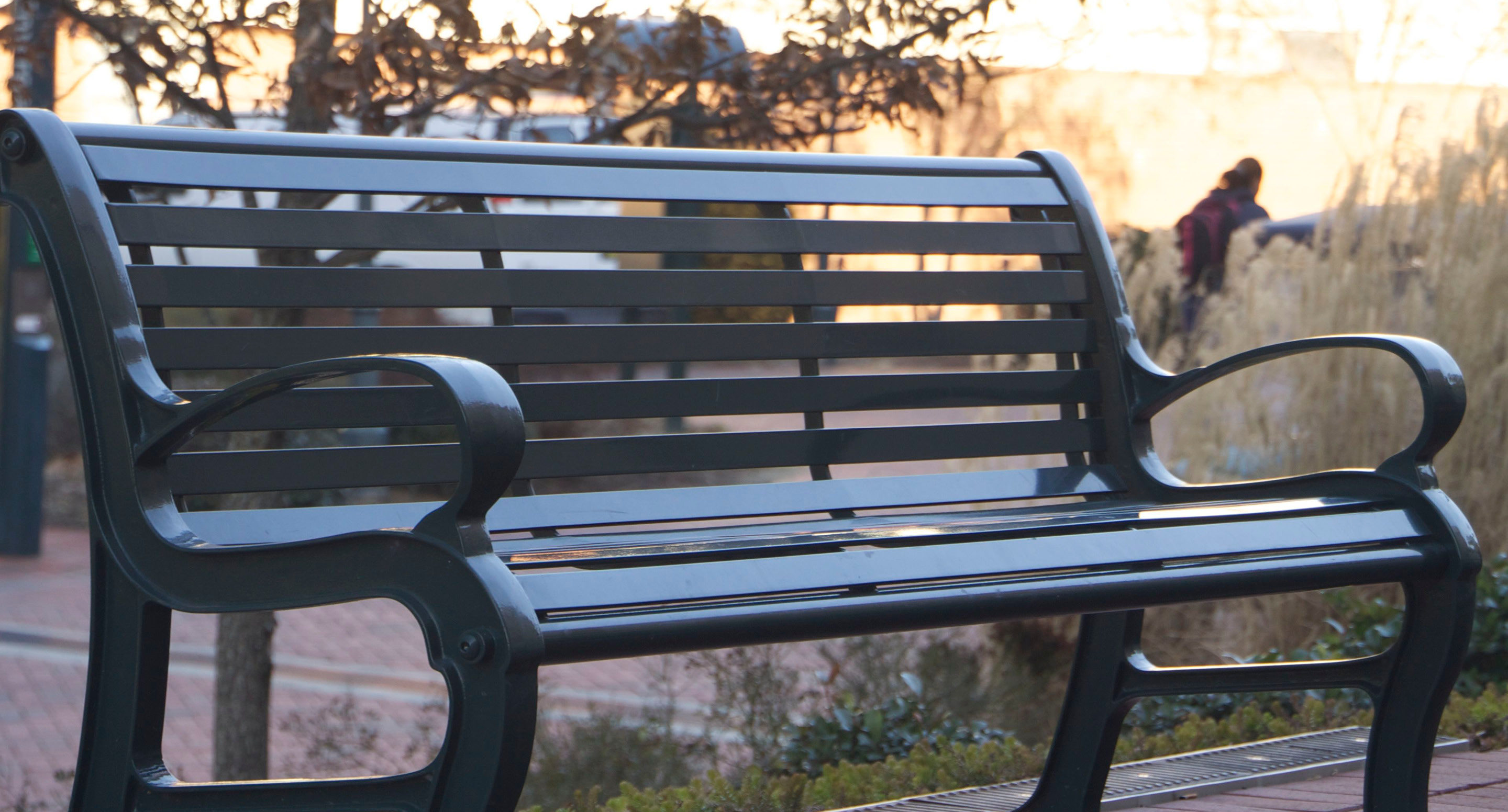 A black metal bench is positioned outdoors on a paved walkway, with a blurred pedestrian and ornamental grasses in the background.