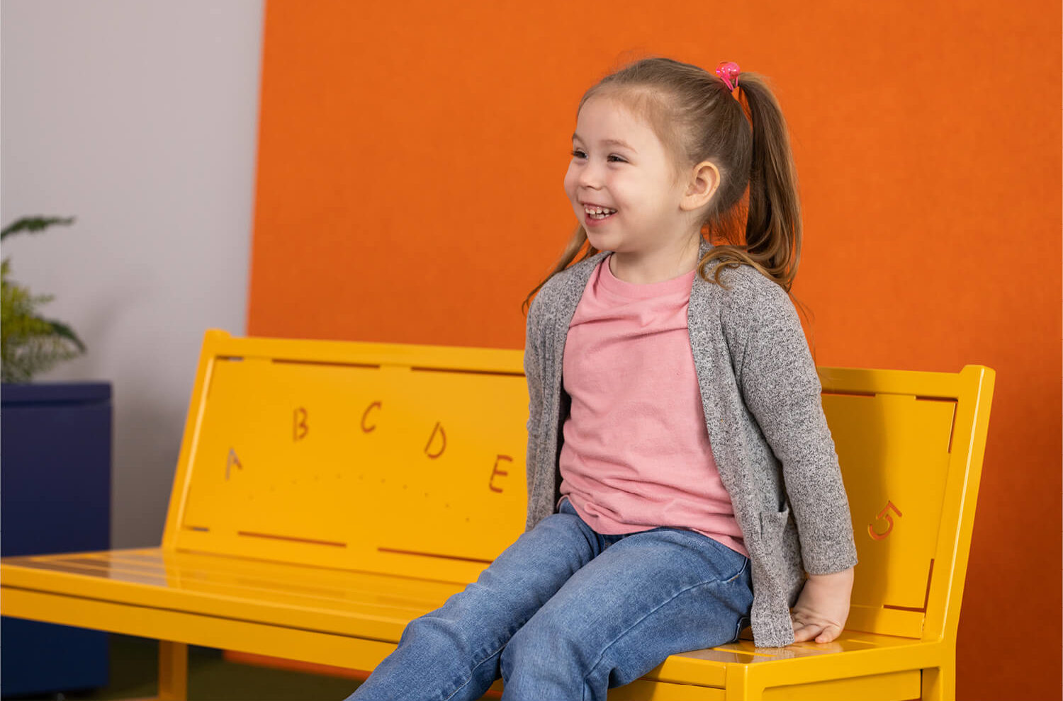 Young girl with pigtails sits on a yellow bench marked with alphabet letters, smiling, wearing a pink shirt, gray cardigan, and jeans. Orange background.