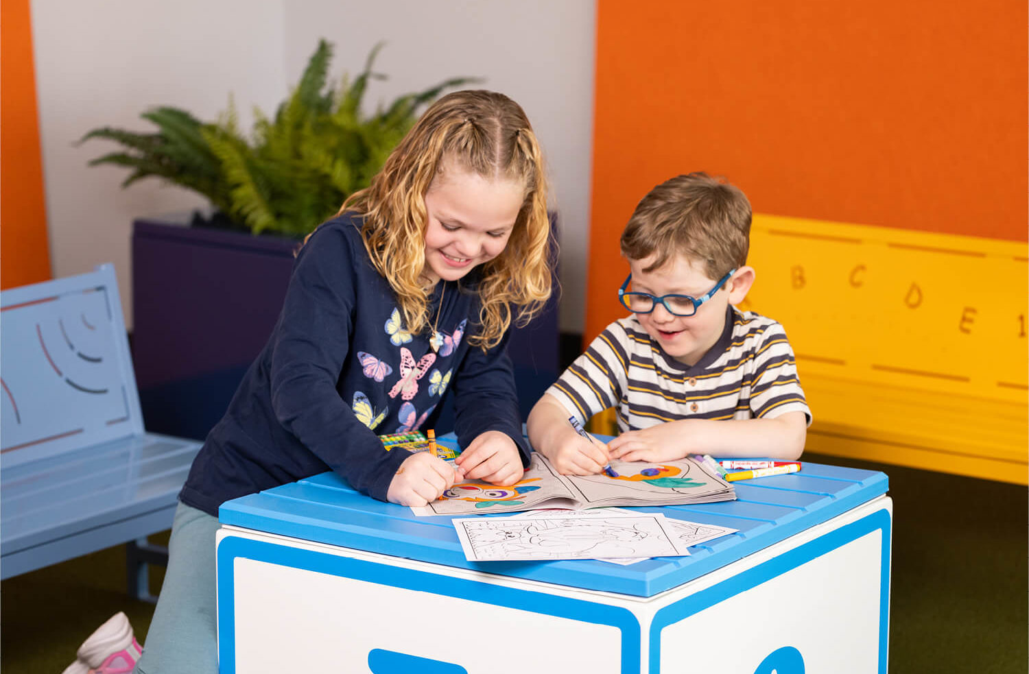 Two young children sit at a blue table and color in activity books with crayons; there are benches and plants in the background.