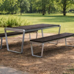 The Ogden Harvest Table with two benches is set on mulch in a park, surrounded by grass and trees.