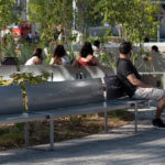 A man sits alone on a metal bench in a park while three women sit together on another bench nearby, surrounded by greenery and paved walkways.