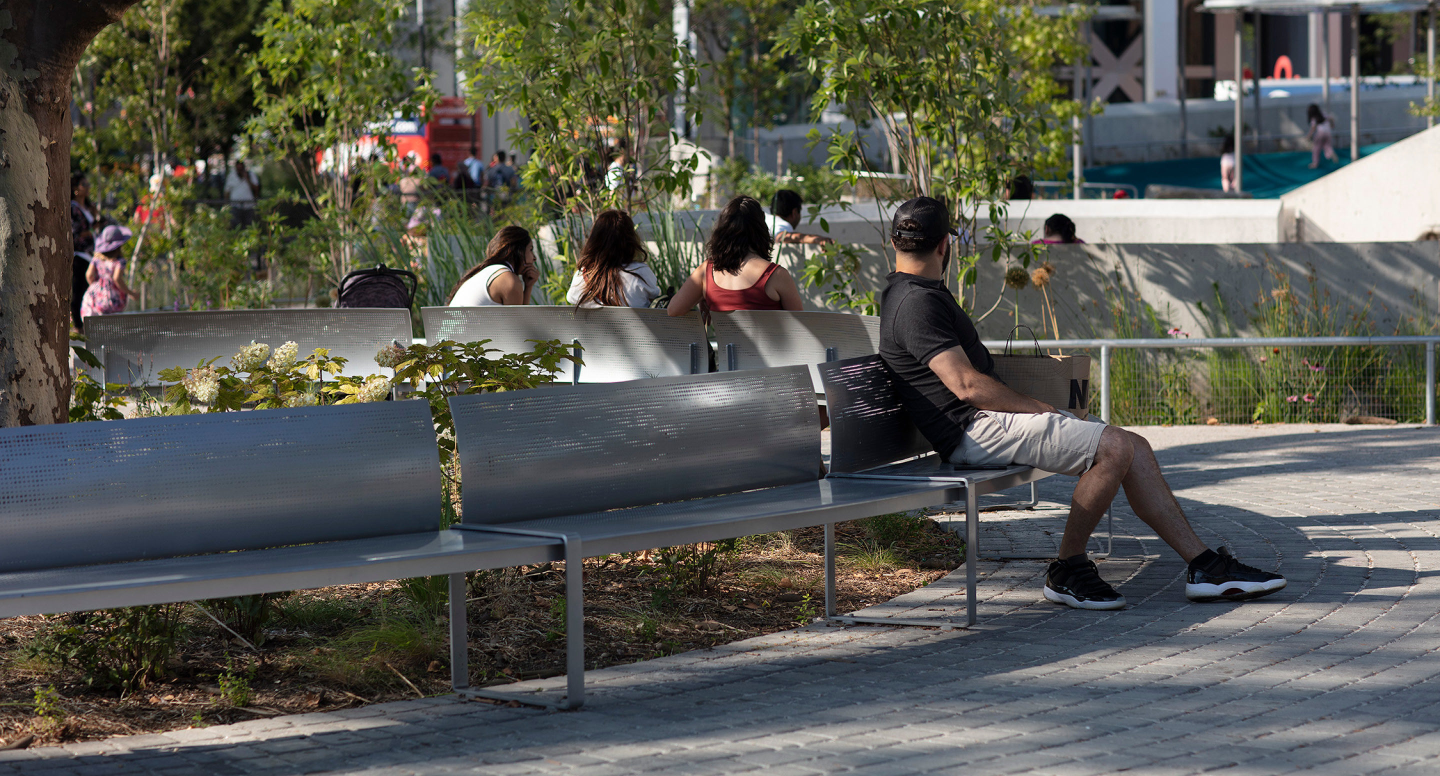 maglin-products_custom-battery A man sits alone on a metal bench in a park while three women sit together on another bench nearby, surrounded by greenery and paved walkways.