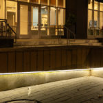 Wooden bench with built-in lighting at the base, situated on a sidewalk at night in front of a building entrance.