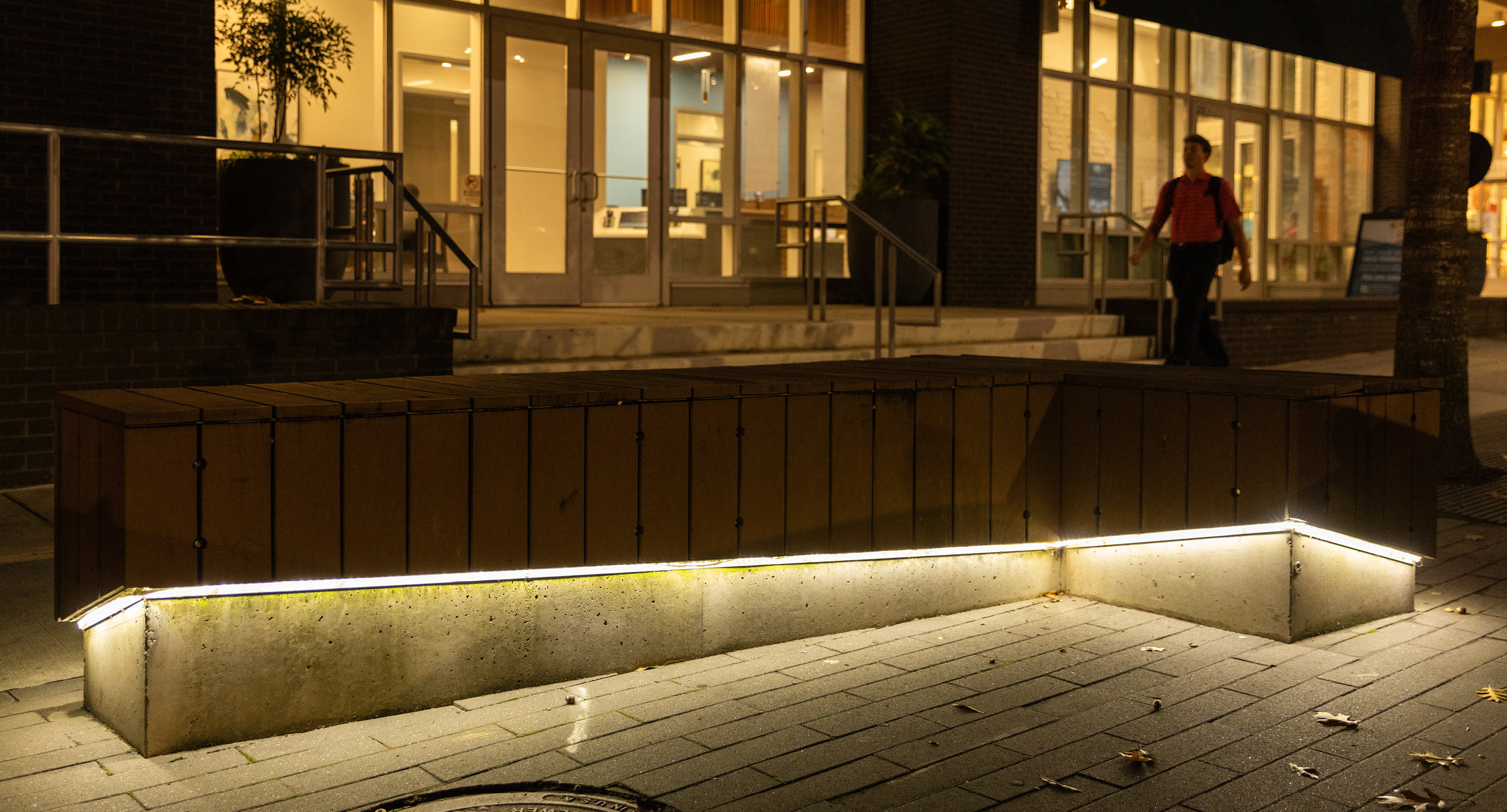 maglin-products_custom-stonewall Wooden bench with built-in lighting at the base, situated on a sidewalk at night in front of a building entrance.