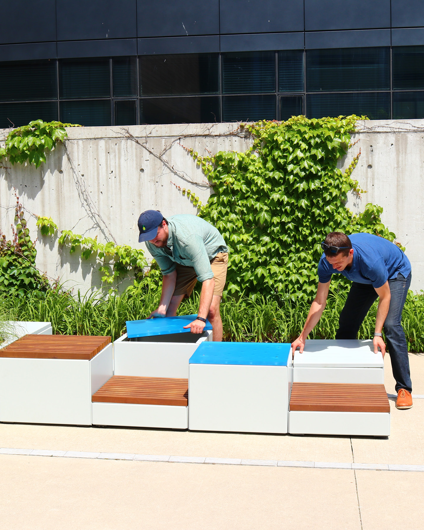 Two men outdoors lift blue lids on modern white benches with wood seating, next to a concrete wall with green vines growing on it.
