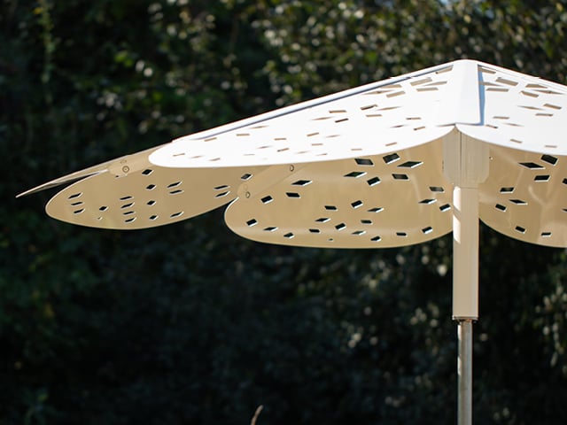 Close-up of a modern, cream-colored outdoor umbrella with leaf-shaped panels featuring geometric cutouts, set against a backdrop of trees.