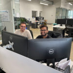 Two men sitting at computers in an office setting, smiling