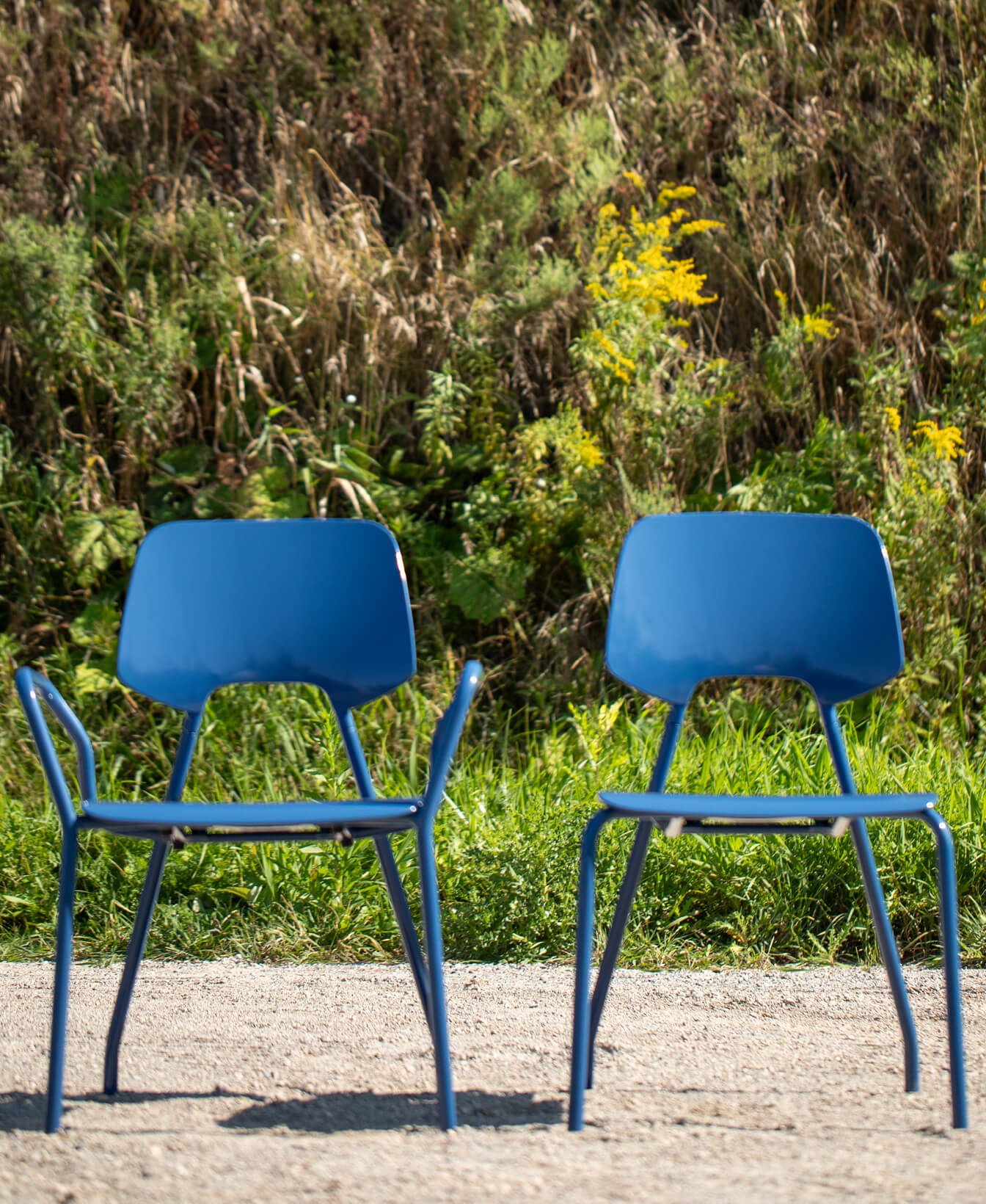 Two ARIES Café Chairs in blue are outdoors on a gravel surface, with green grass and vegetation in the background.