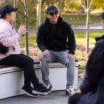 Four people sit and talk outdoors on a curved white bench near orange flowers, wearing casual jackets and caps on a sunny day.