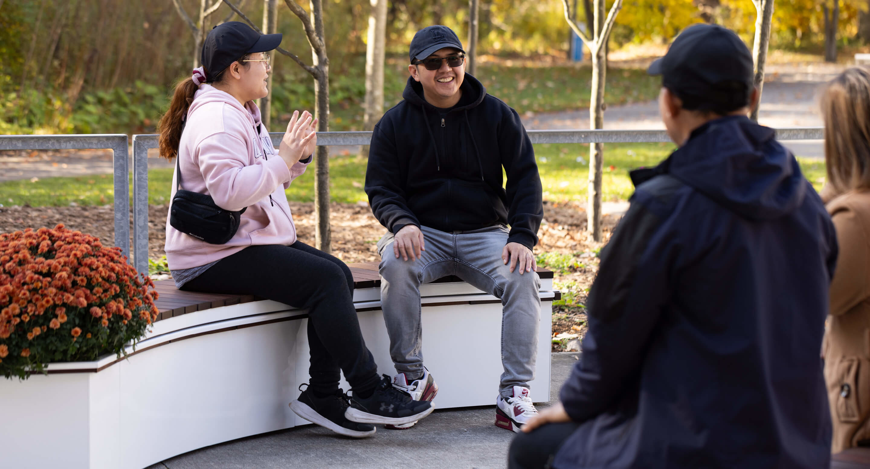 maglin_canoe-planters_2 Four people sit and talk outdoors on a curved white bench near orange flowers, wearing casual jackets and caps on a sunny day.