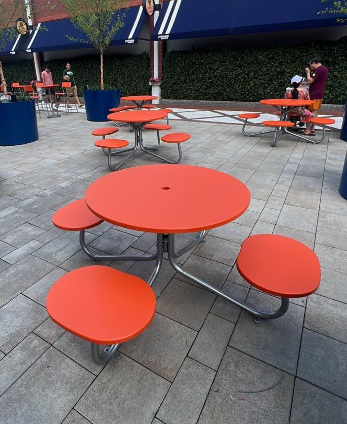 Several FAVA Cluster Seating units—round orange picnic tables with attached seats—are set up on a paved outdoor area, with people visible in the background near planters and greenery.