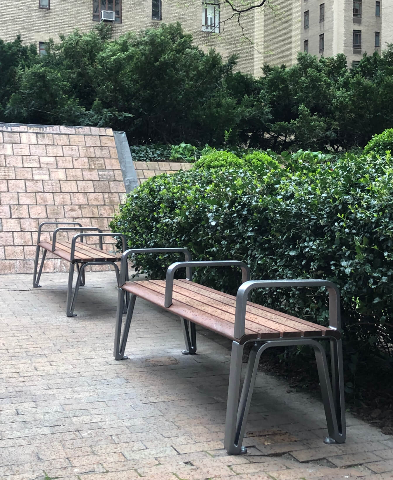 Two Iconic Backless Benches with metal frames are set on a brick walkway beside green bushes in an outdoor urban space.