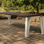 A modern wooden bench with white metal legs sits on a paved area outdoors, with trees and rocks in the background.
