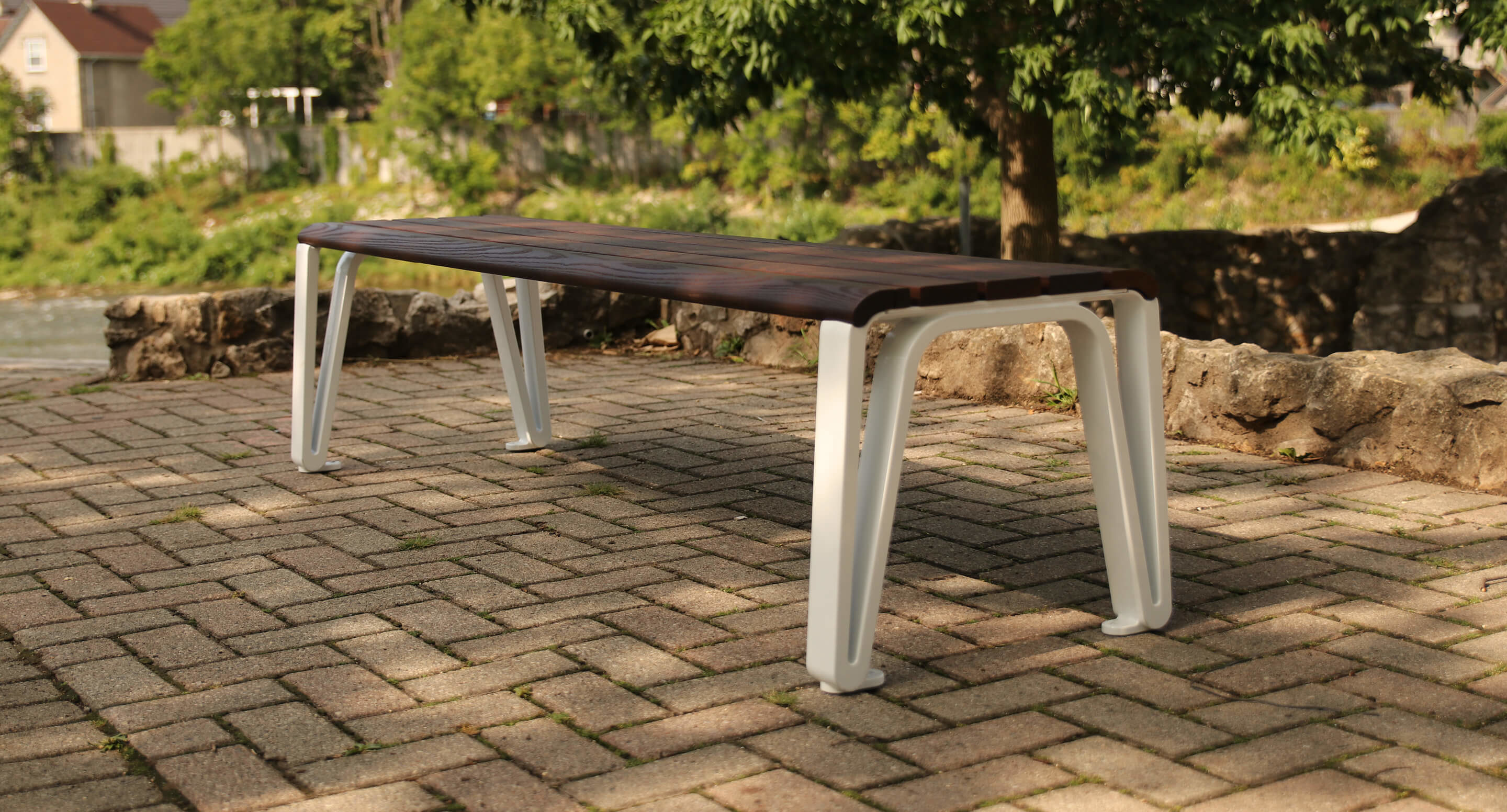 maglin_iconic-backless-bench_1 A modern wooden bench with white metal legs sits on a paved area outdoors, with trees and rocks in the background.