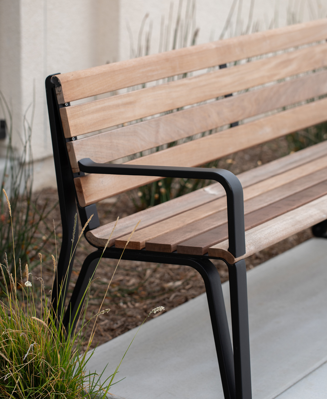 The Iconic Backed Bench, featuring a modern wooden seat and black metal frame, is placed on a concrete surface near ornamental grass and a building wall.