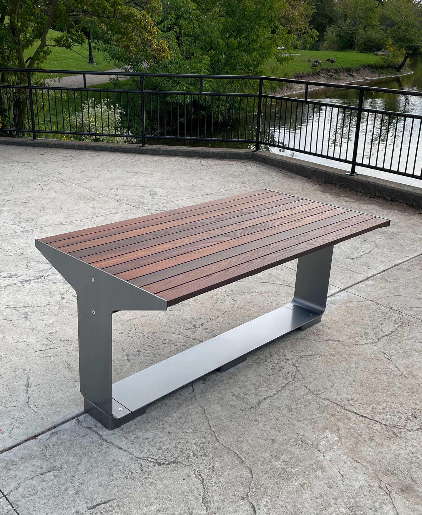 An L-Series Table, featuring a metal base and wooden slats, is set on a concrete surface near a black metal fence and a pond.