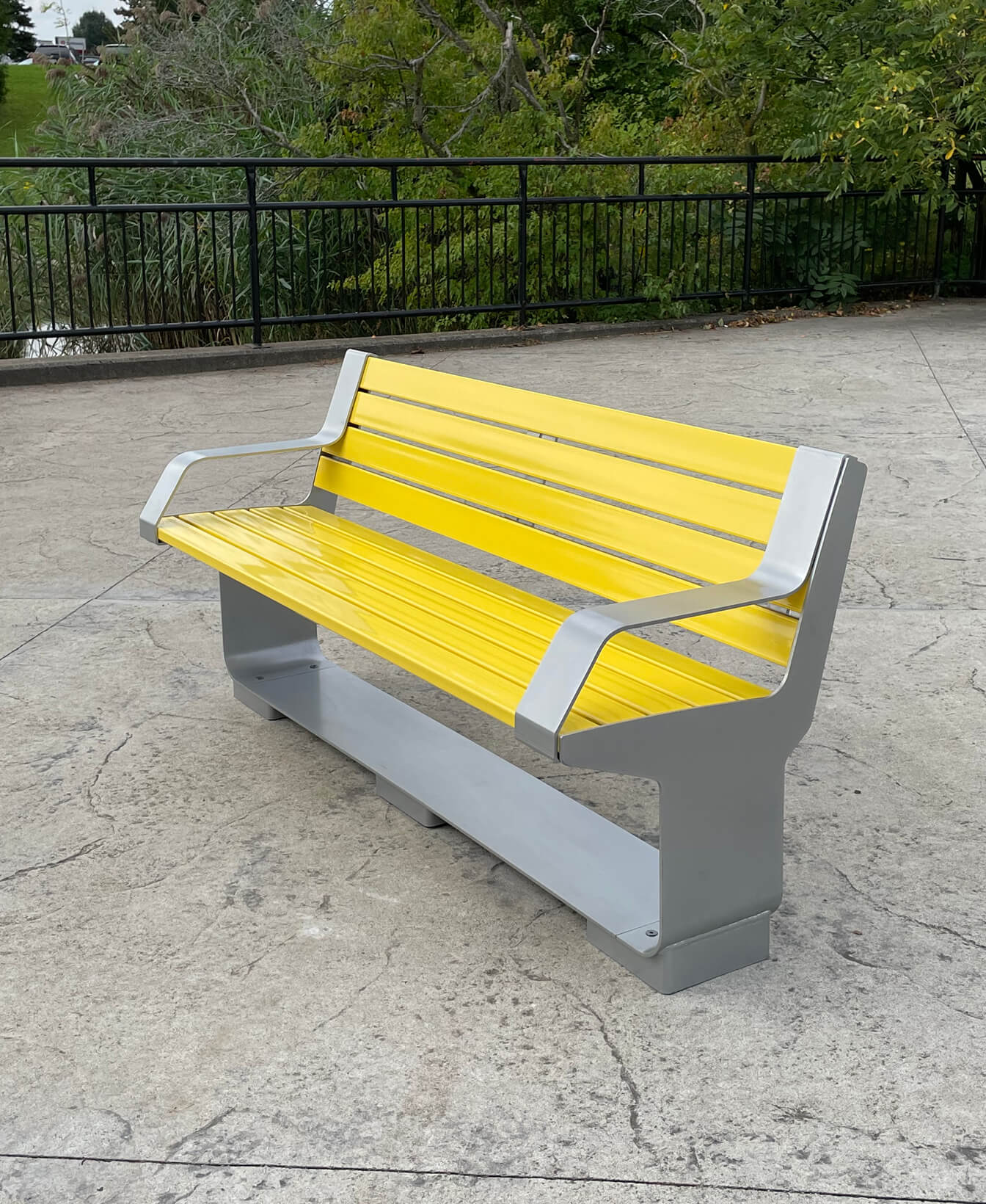 An L-Series Backed Bench in yellow and gray is placed on a concrete surface near a black metal fence, with greenery visible in the background.