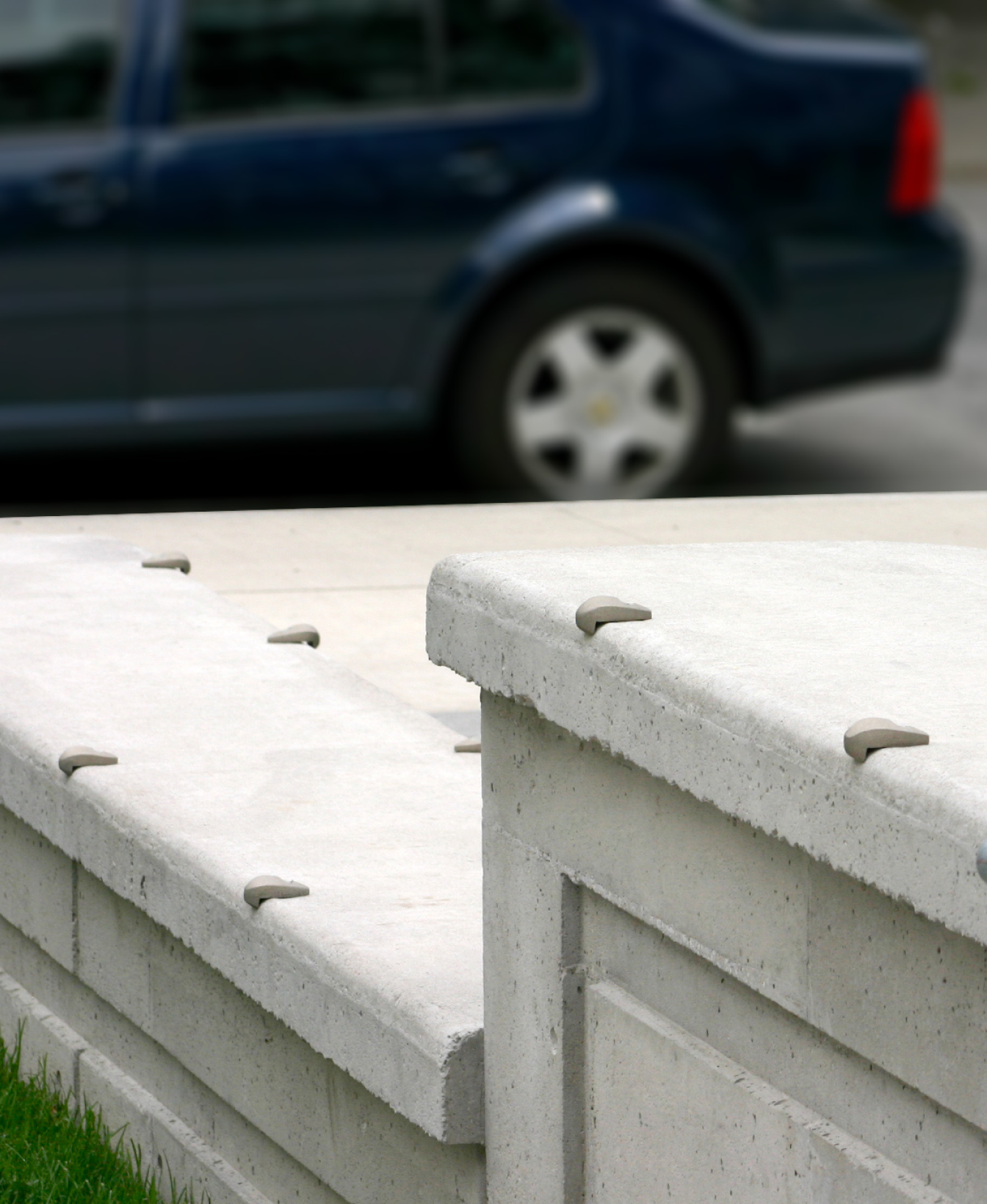A 100 Skate Deterrent - Wall Mount with small, rounded metal spikes is installed atop a concrete wall, with a blue car visible on the street in the blurred background.
