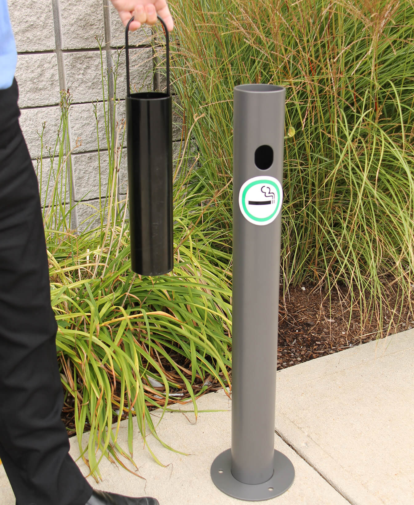 A person removes the cylindrical container from a 300 Ash Receptacle, a gray outdoor cigarette disposal unit located near a sidewalk and plants.