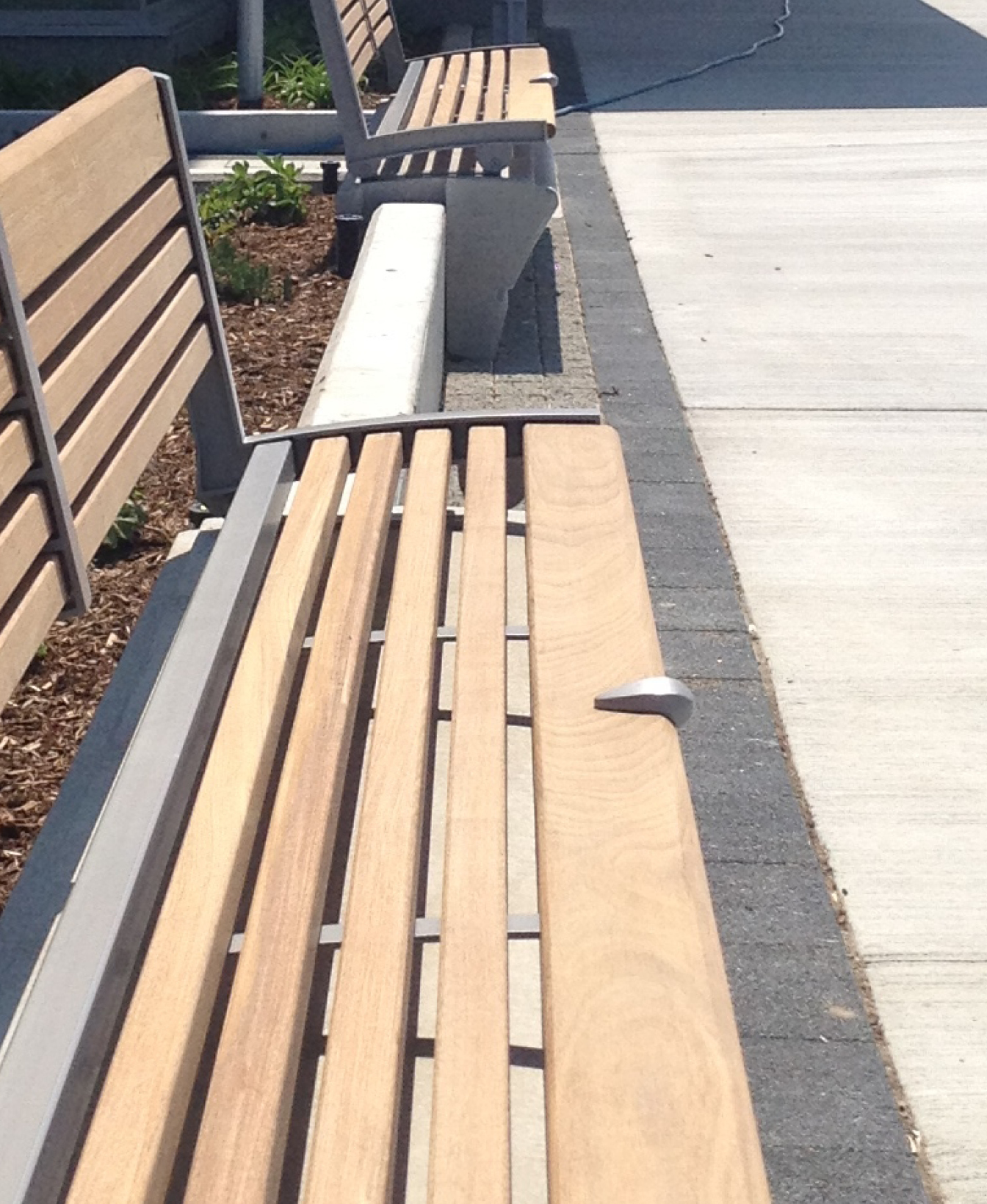 A 100 Skate Deterrent - Bench Mount is installed on an outdoor wooden bench with metal armrests beside a sidewalk; a white plastic spoon rests on the seat.