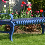 A blue metal bench sits on green grass in front of a flower bed with blooming red, white, and pink tulips.