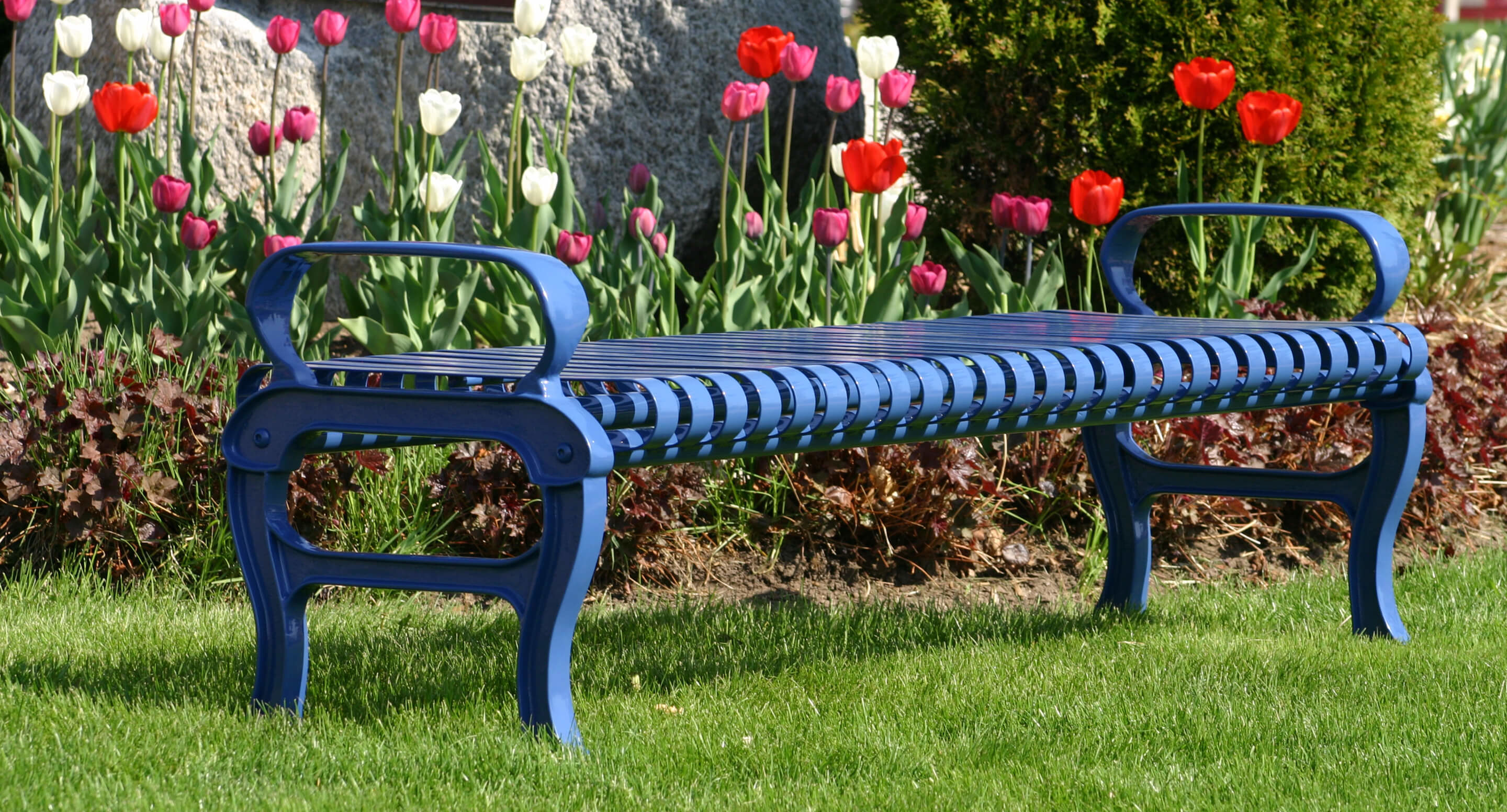 maglin_mbe-0300-backless-bench_1 A blue metal bench sits on green grass in front of a flower bed with blooming red, white, and pink tulips.