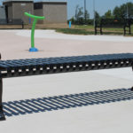 A black metal park bench on a concrete surface with other benches, a green water feature, grass, and buildings in the background.