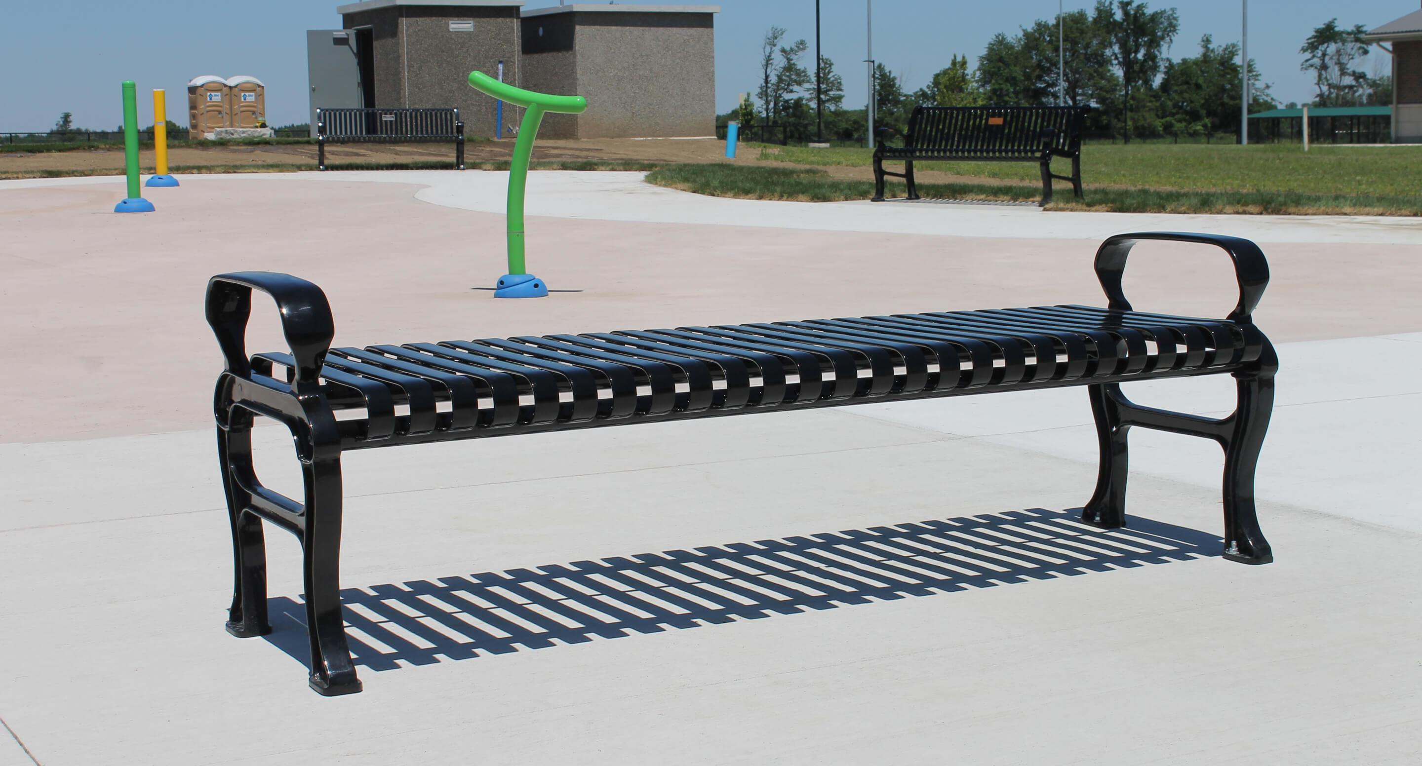 A black metal park bench on a concrete surface with other benches, a green water feature, grass, and buildings in the background.