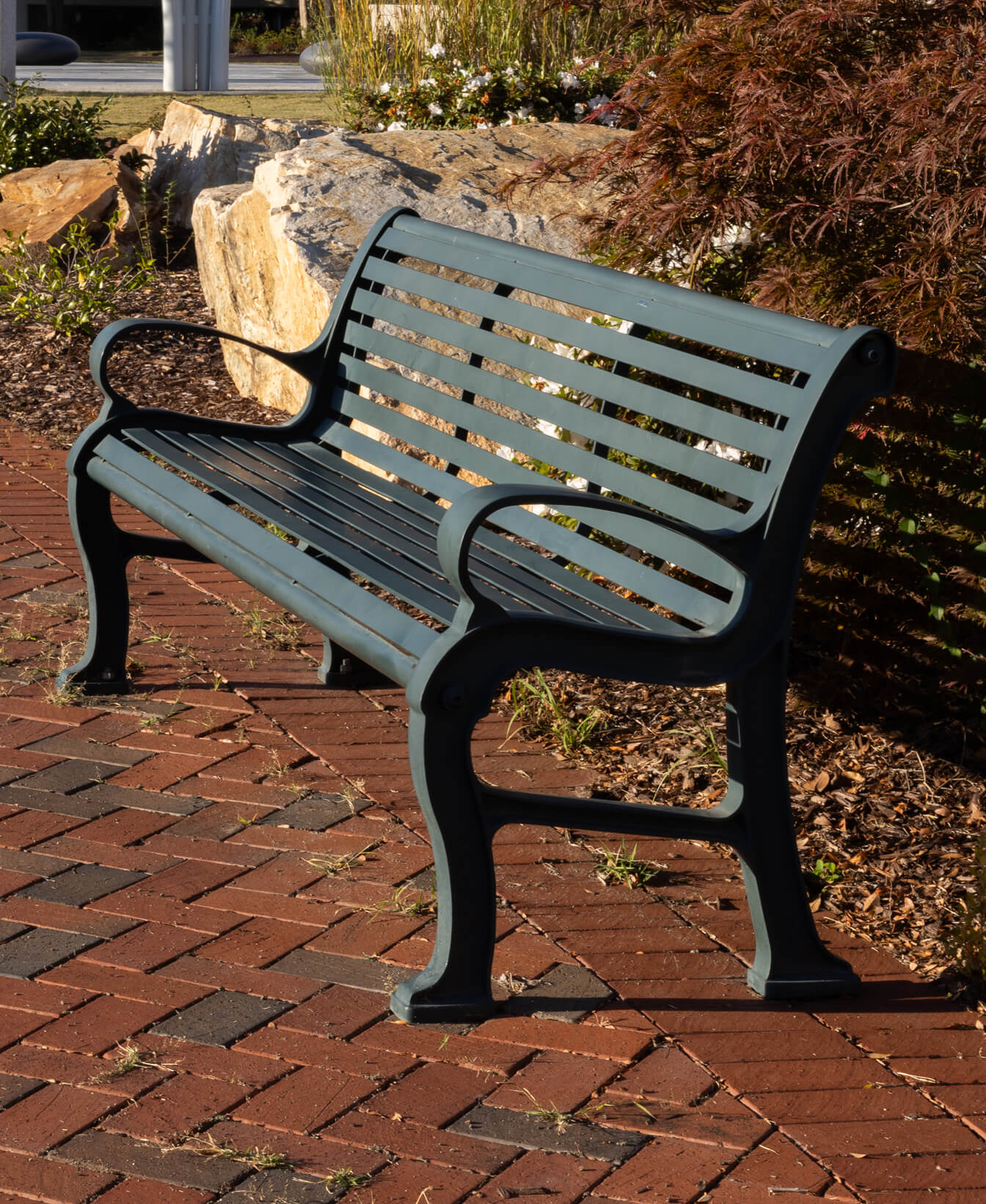 The 300 Backed Bench, a metal bench with horizontal slats, sits on a brick walkway surrounded by plants and rocks in an outdoor setting.