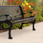 A black metal bench sits on a concrete sidewalk next to a planted area with green leaves and orange and yellow flowers.