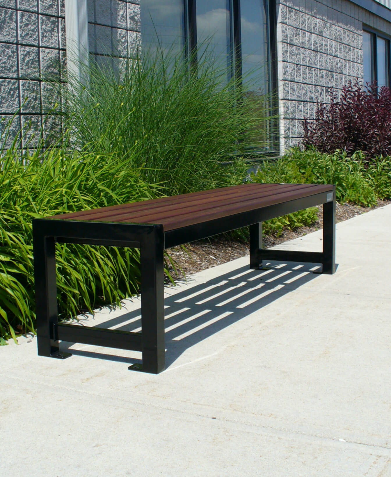 The 400 Backless Bench features a dark wooden seat and black metal frame, positioned on a concrete sidewalk near a building and lush green plants for a modern outdoor setting.