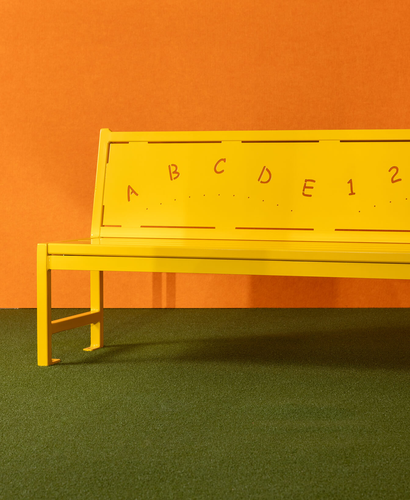 Le Banc 400 pour enfants, un banc en métal jaune avec des lettres et des chiffres découpés dans le dossier, est placé contre un mur orange sur un sol vert.